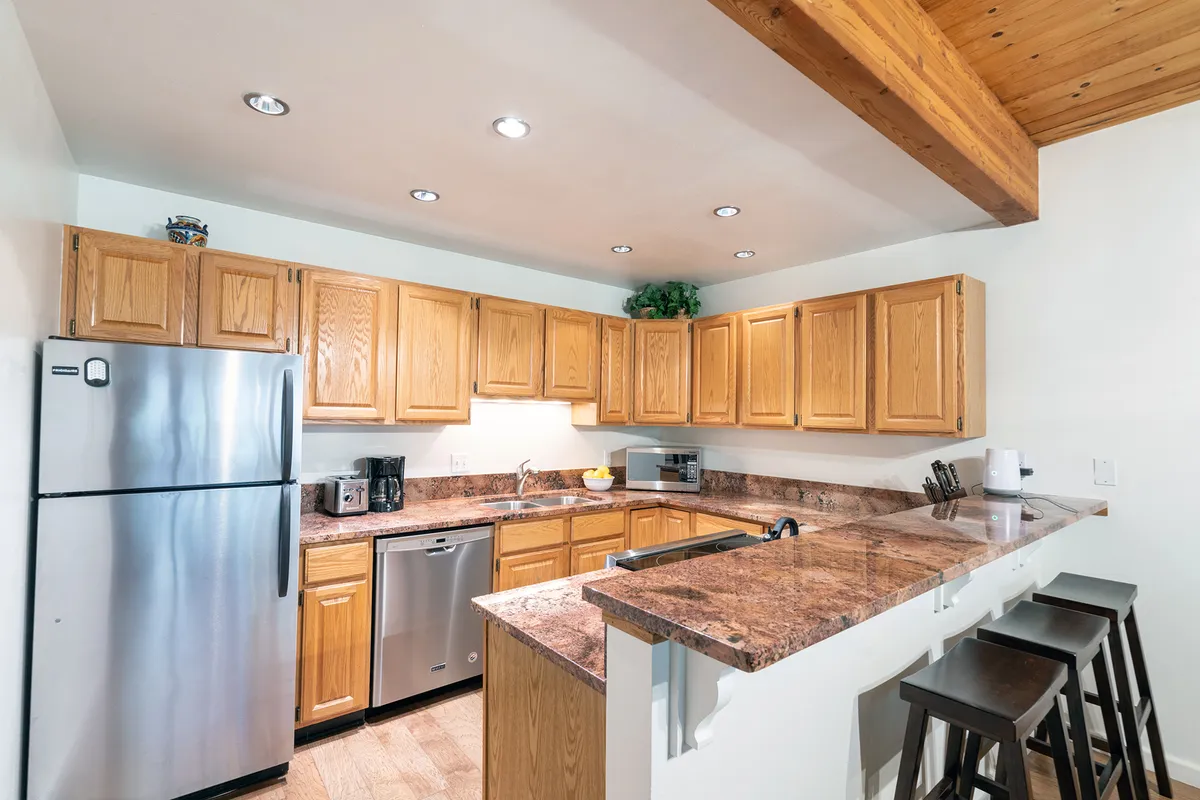 A spacious, recently updated kitchen featuring stainless steel appliances, granite countertops, wooden cabinetry, and a bar seating area. The kitchen is open to the living and dining areas, inviting guests to cook, converse, and entertain in comfort.