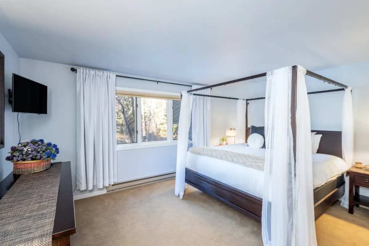 A calm, inviting bedroom with a four-poster bed, large window letting in natural light, a television, and decorative touches like a basket of flowers. The space exudes warmth and comfort, ideal for families with young children or anyone seeking relaxation after arrival.