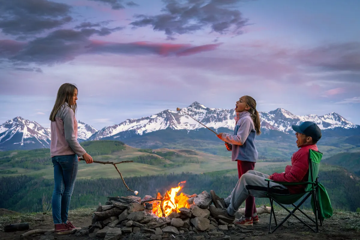 Children gather around a glowing campfire, roasting marshmallows as the sun sets behind the mountains. Adults are nearby, creating a warm and nostalgic atmosphere filled with laughter and storytelling.