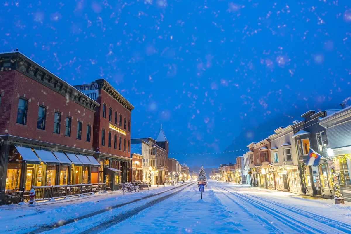 This image depicts Telluride's snow-covered Main Street, lined with historic buildings and festively decorated trees. Pedestrians walk along the charming, bustling street, hinting at a vibrant local scene ideal for holiday shopping, dining, and exploration. The magical ambiance suggests a lively, walkable mountain town atmosphere.