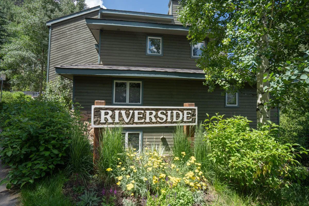 A picturesque scene featuring the 'RIVERSIDE' wooden sign, vibrant greenery, and colorful flowers, set by the river. The natural setting hints at lovely outdoor experiences like hiking and family gatherings.