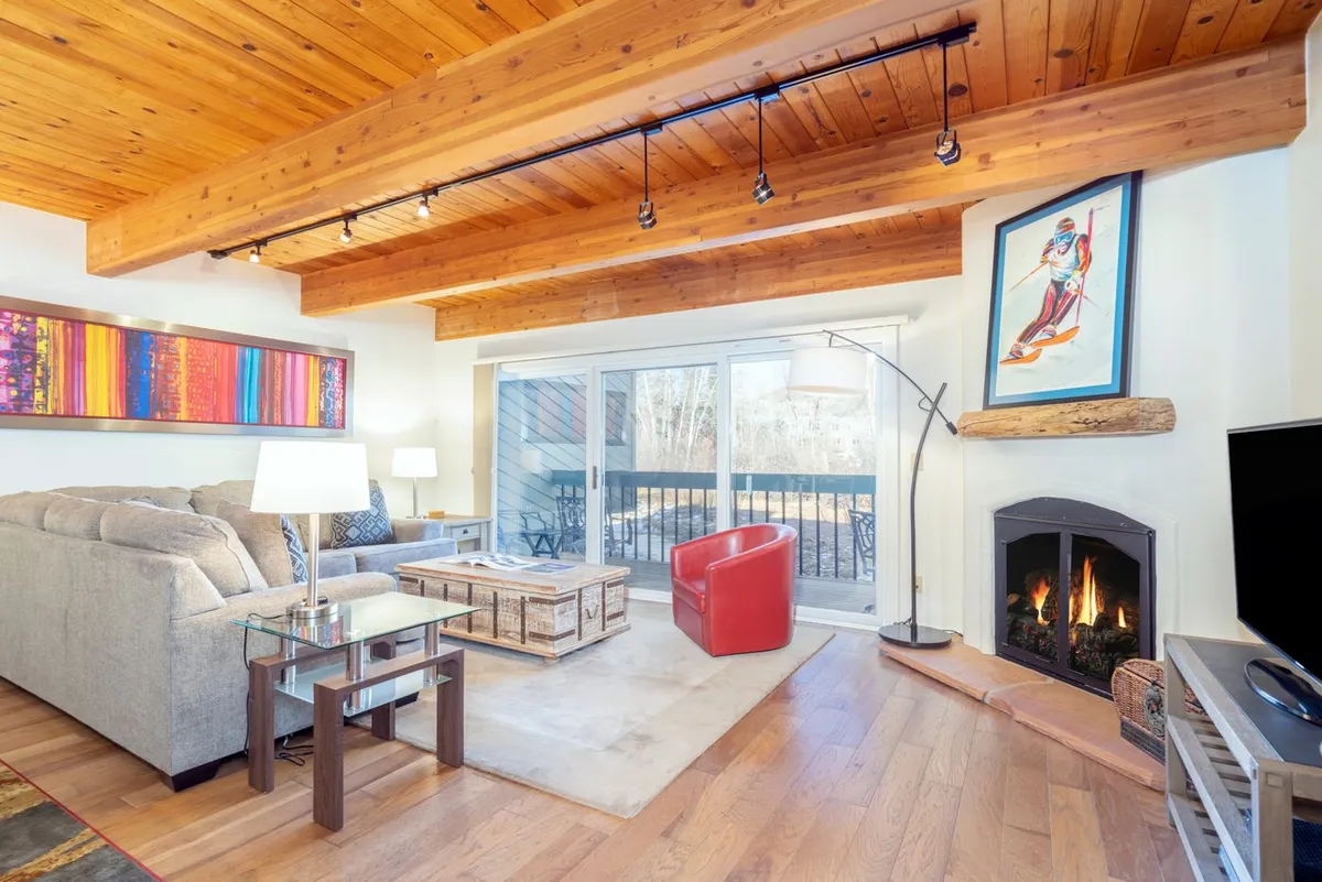 A cozy living room with a plush sofa, inviting red chair, and a warm fireplace as the focal point. Large windows offer glimpses of the landscape beyond, while a spacious coffee table stands ready for games or gatherings. Tasteful artwork adds a touch of mountain charm to the bright, open space.