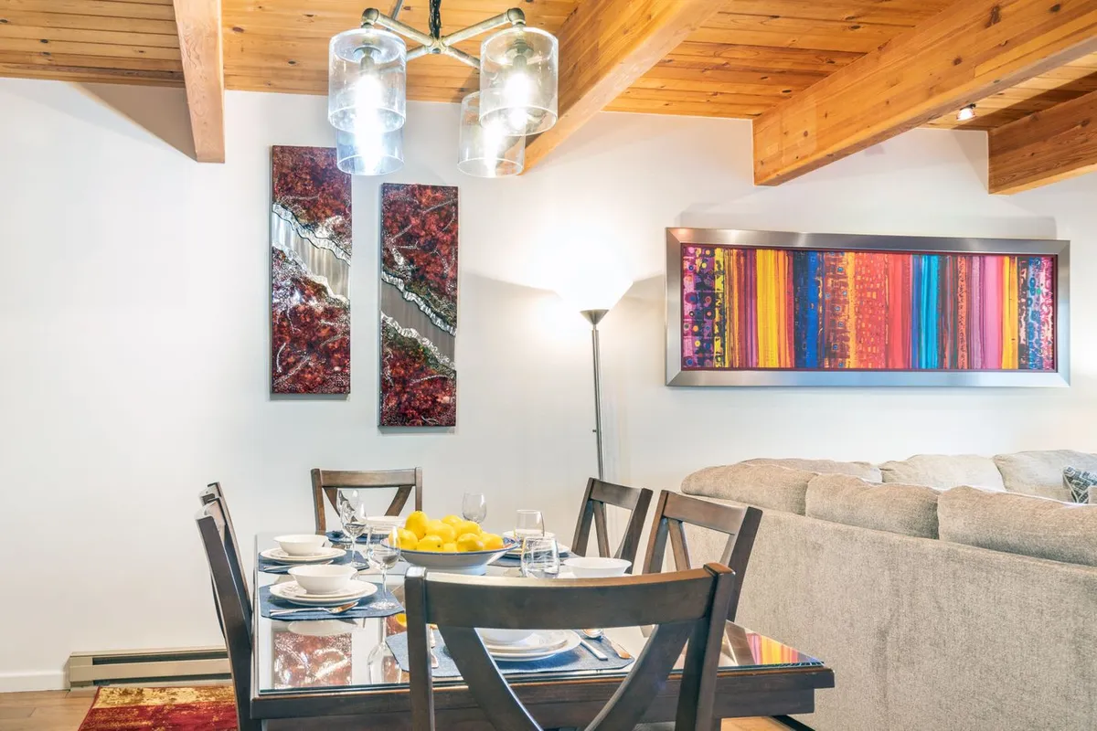 A welcoming dining area with a table set for a meal, surrounded by comfortable seating and vibrant abstract wall art. The adjoining sofa and cushions hint at the seamless connection between dining and living spaces.