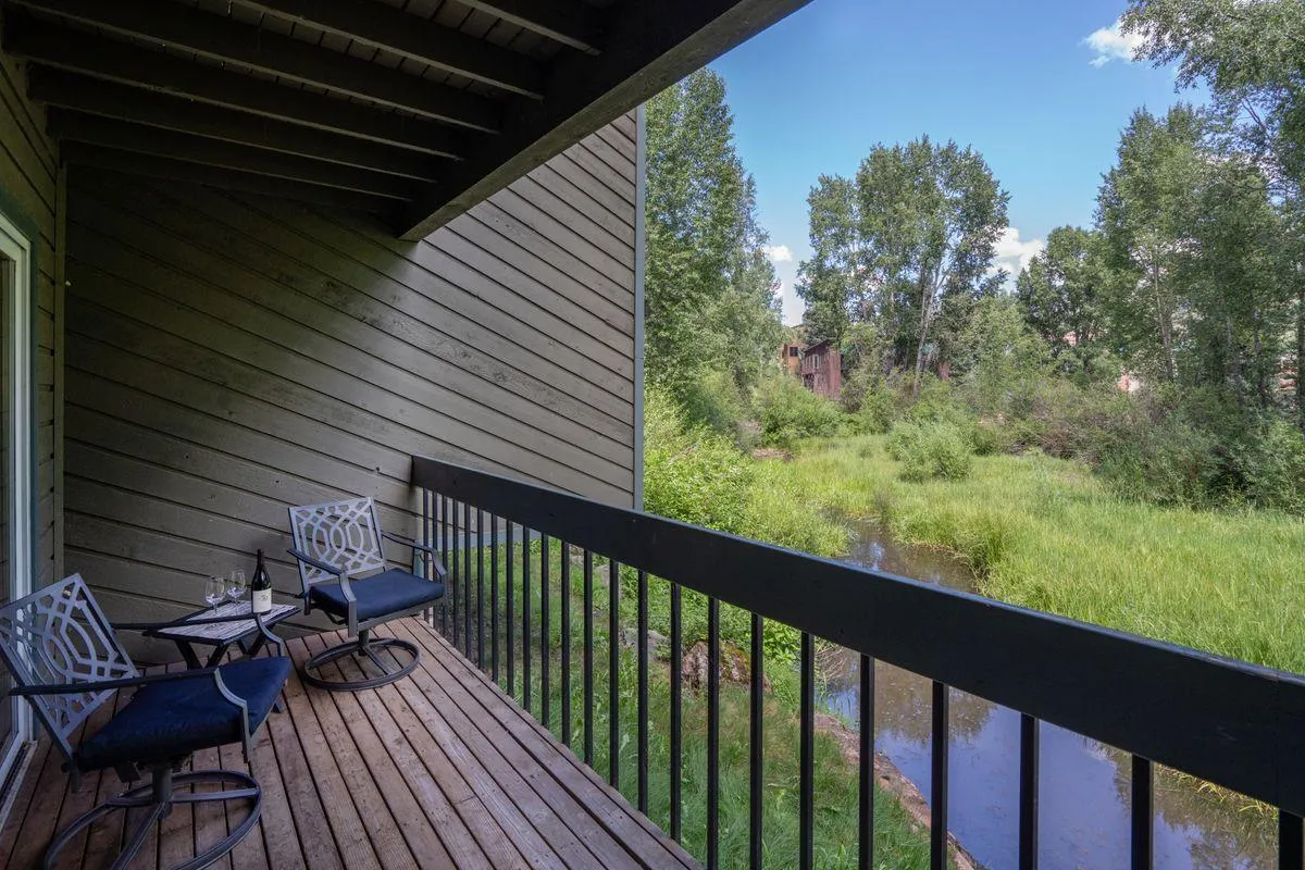 An inviting exterior deck or balcony setup with comfortable seating, wine glasses on the table, and a tranquil view overlooking a creek or river. The natural surroundings suggest peaceful mornings and relaxing moments spent outdoors.
