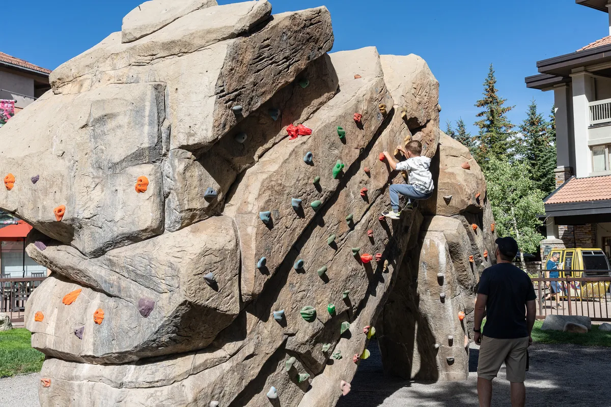 A young child is climbing a colorful outdoor climbing wall while an adult closely supervises, set against a backdrop of greenery and play structures. The scene captures active engagement, encouragement, and family bonding during an outdoor adventure.