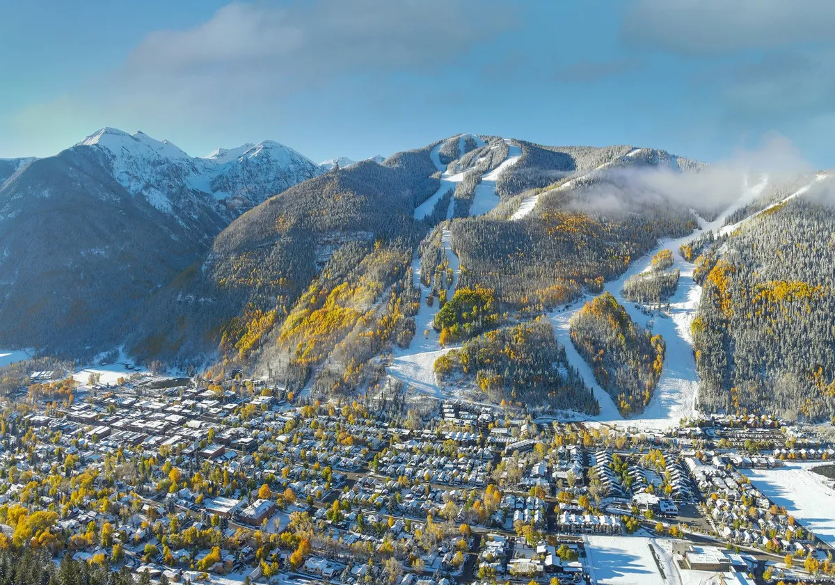 A sweeping exterior shot of snow-covered mountains and ski slopes, vibrant autumn trees, and the town of Telluride nestled below. The scene marries both winter and autumn, showcasing the area’s year-round appeal for hiking, skiing, and scenic exploration.
