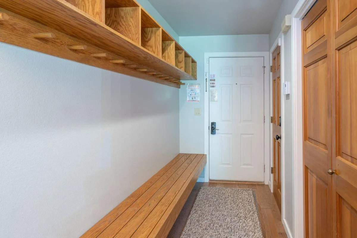 This image shows a welcoming entryway featuring a sturdy wooden bench and open shelves for storage. The front door is visible, and the setup is clearly designed to help guests transition smoothly from outdoors to indoors, offering space to sit, remove shoes, and organize gear.