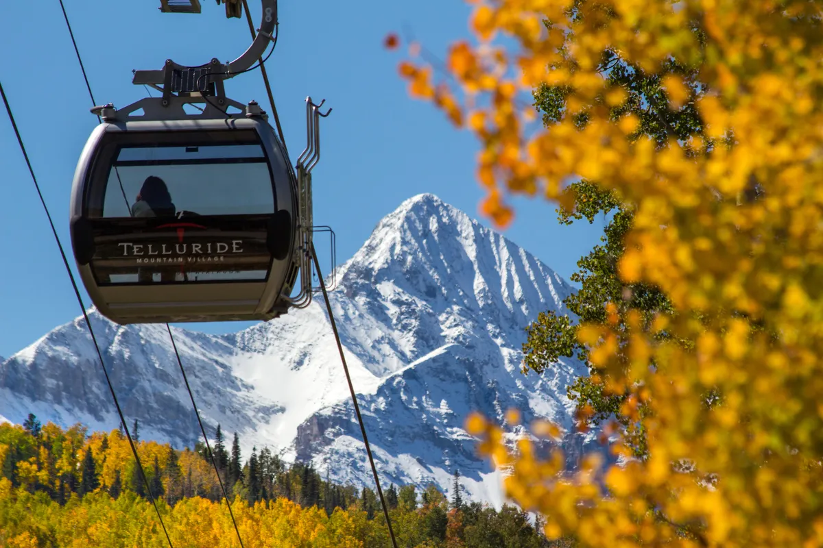 A scenic view featuring the Telluride gondola gliding above autumn foliage, with a snow-capped mountain rising in the background. The image captures the essence of Telluride's outdoor adventure spirit, showcasing easy access to world-class skiing and breathtaking natural beauty.
