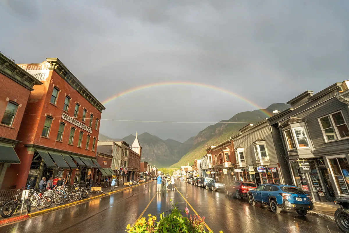 A lively street view of historic Telluride, with colorful buildings, a rainbow over shiny wet streets, pedestrians strolling, and bikes parked along the sidewalk. The scene captures the vibrant local atmosphere and community spirit.