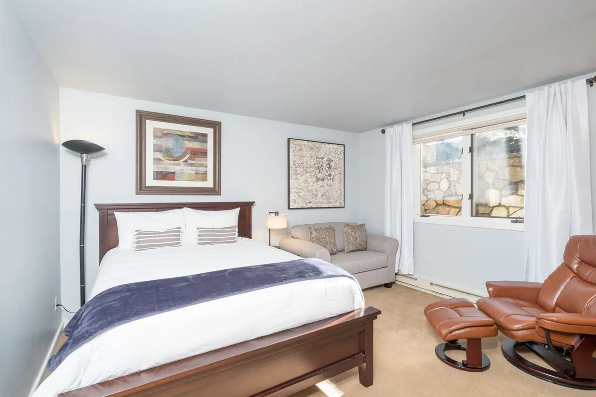A serene bedroom featuring a comfortable bed adorned with plush bedding, a soft armchair for reading or relaxation, tasteful artwork on the wall, and a large window inviting in abundant natural light. The overall design radiates tranquility, making it an ideal space for restful nights and lazy mornings.